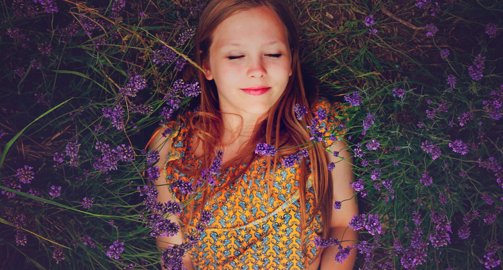 Keltaiseen mekkoon pukeutunut lapsi makaa violettien kukkien keskellä ja hymyilee silmät kiinni.