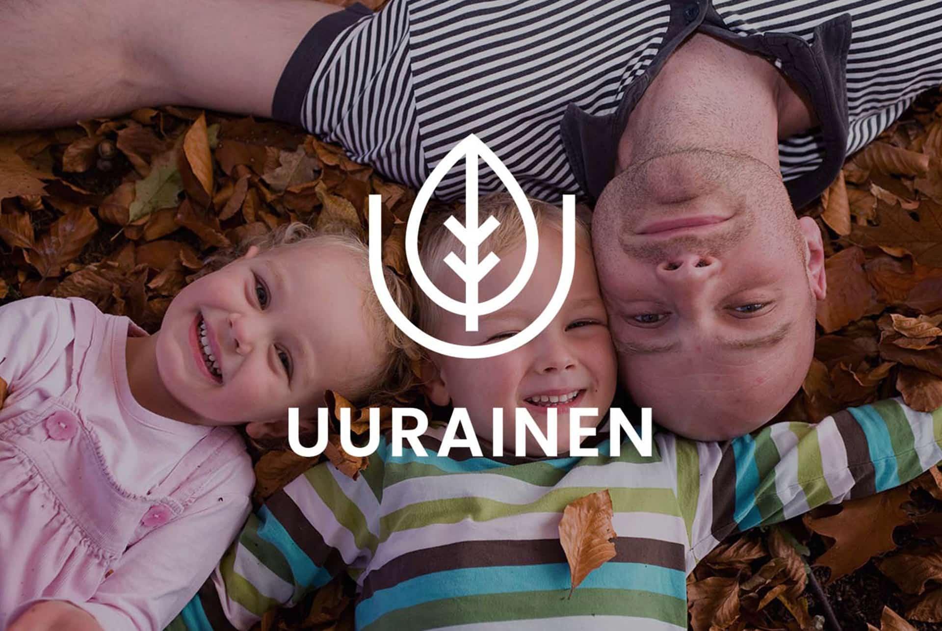 Kaksi lasta ja aikuinen makaavat lehtien päällä maassa ja hymyilevät kameralle, päällä Uuraisten kunnan logo.